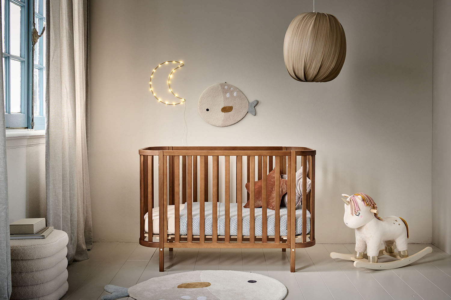 Lit bébé ovale en bois couleur noyer, installé dans une chambre douce et naturelle avec décoration murale en forme de lune et poisson, et un cheval à bascule licorne.